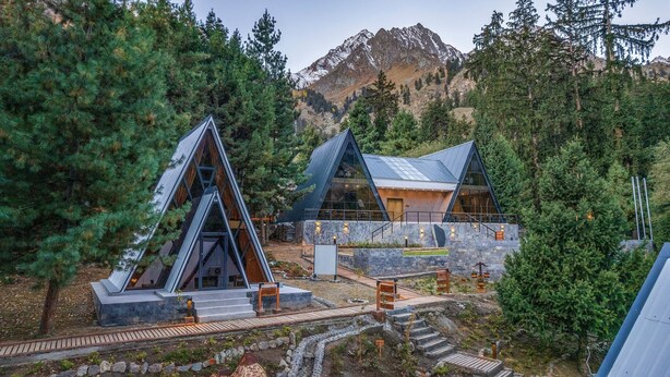 A modern mountain lodge with triangular roofs and stone walls, surrounded by dense forest and mountains.