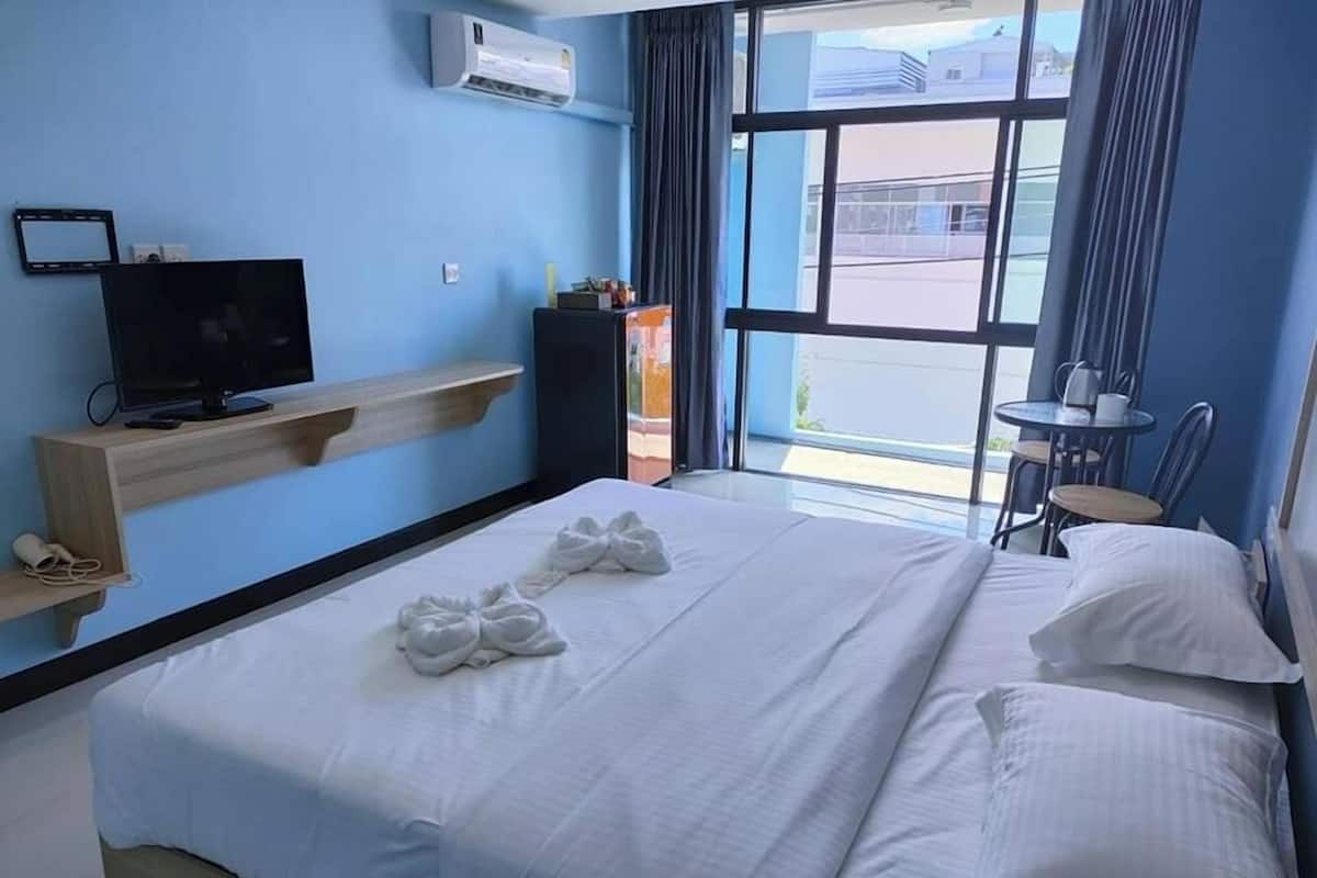 A hotel room with a bed, a TV on a wooden shelf, a small table with a chair, and a window with curtains.