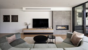 A modern living room with a large L-shaped sofa, a flat-screen TV, a fireplace, and a minimalist design. - Bayside Grandeur with Heated Pool, Spa and Sauna plus each Bedroom has Ensuite (Bentleigh)