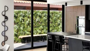 A modern kitchen with a large island, bar stools, and a view of lush greenery outside. - Bayside Grandeur with Heated Pool, Spa and Sauna plus each Bedroom has Ensuite (Bentleigh)