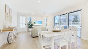 Een lichte, ruime kamer met een eettafel, stoelen, een bank en een groot raam met uitzicht op de omgeving.
