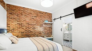 Une chambre avec un mur en briques, un lit avec une literie à rayures, une table de chevet, un luminaire suspendu et un téléviseur fixé au mur.