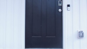 A black door with a keypad lock, a doormat with "WELCOME HOME" text, and two wall-mounted lanterns.