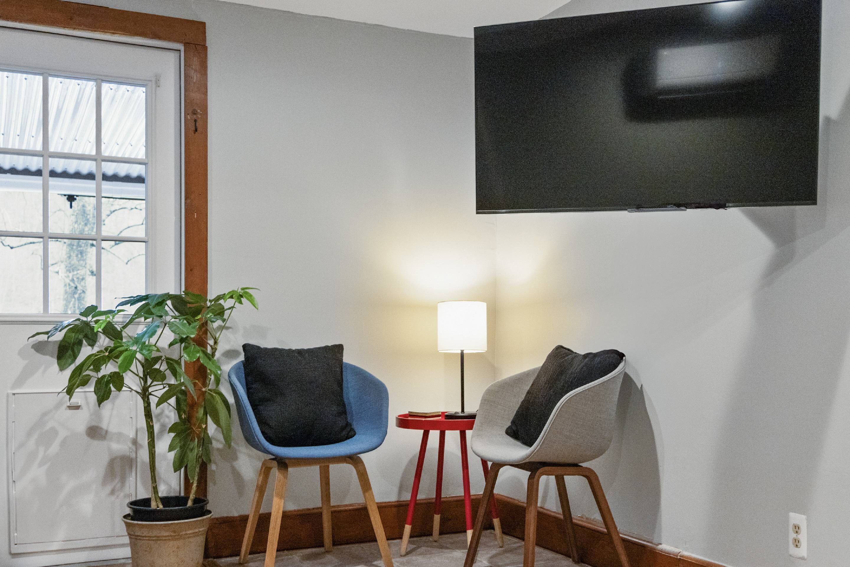 Une pièce avec un téléviseur fixé au mur, deux chaises, une petite table avec une lampe et une plante en pot.
