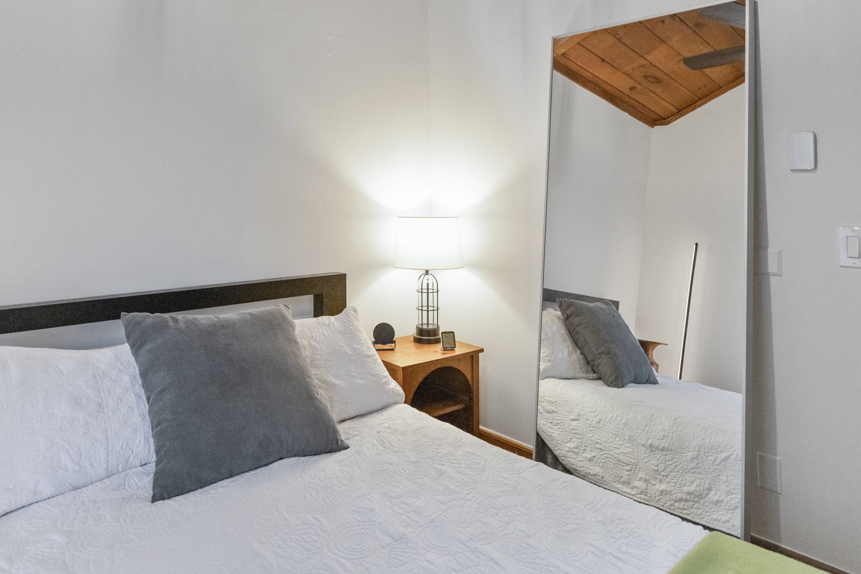 Una cama bien hecha con ropa de cama blanca y gris, una lámpara de noche y una mesita de noche de madera.