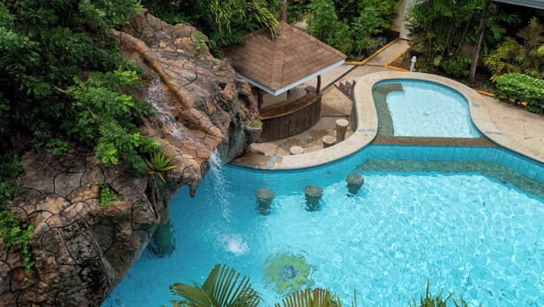 Una piscina con una cascada, rodeada de exuberante vegetación y una pequeña estructura de madera.