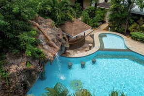 Una piscina con una cascada, rodeada de exuberante vegetación y una pequeña estructura de madera.