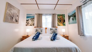 Una habitación de hotel con una cama grande, dos mesitas de noche, una lámpara de techo y un cuadro con un caballo, una playa y un barco.