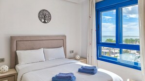 Een net opgemaakt bed met witte lakens en twee blauwe handdoeken, een decoratief object aan de muur, een nachtkastje met een sterrenpatroon en een raam met uitzicht op een strand en gebouwen.