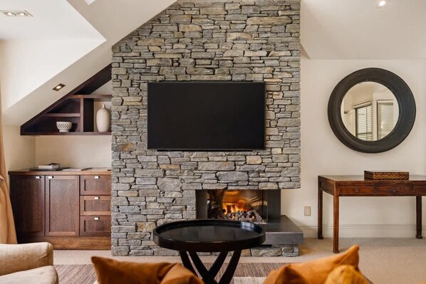 A living room with a stone fireplace, a flat-screen TV mounted above it, a round mirror on the wall, and a wooden table.