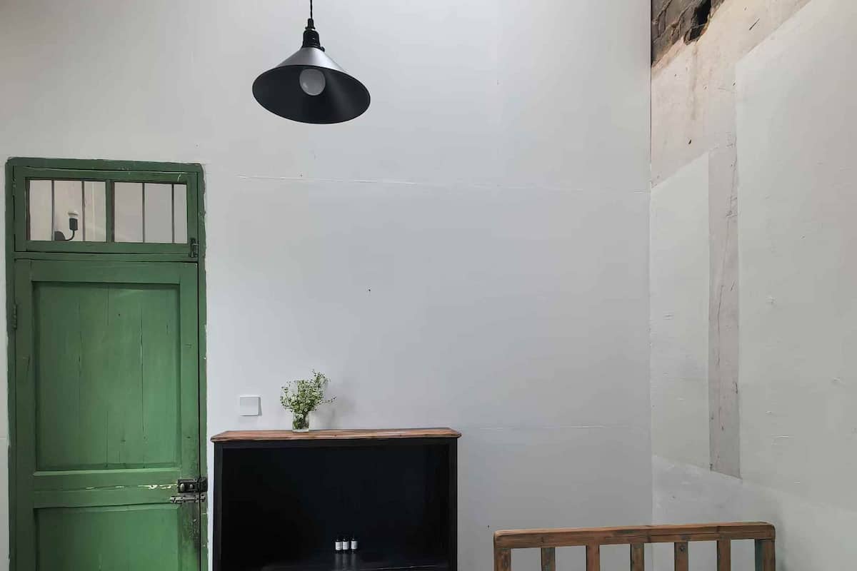 Une petite pièce simple, avec une porte verte, un lit en bois, une petite table de chevet en bois, une lampe suspendue et un mur en briques.