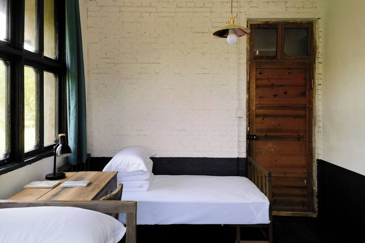 Un lit simple avec une literie blanche, un bureau en bois avec une lampe et une fenêtre avec des rideaux.