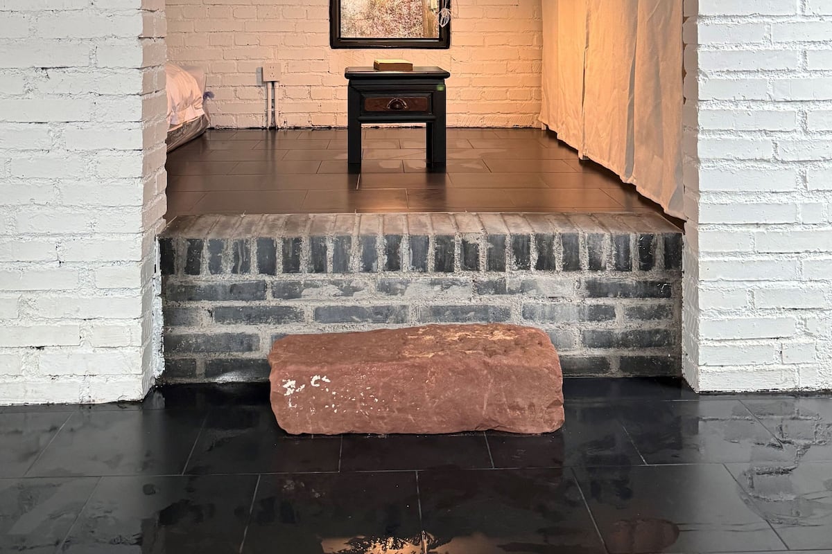 Une pièce aux murs en briques blanches, avec un plafond en bois, un plancher noir, une petite table noire sur laquelle est posée une photo encadrée, et un banc en briques rouges.