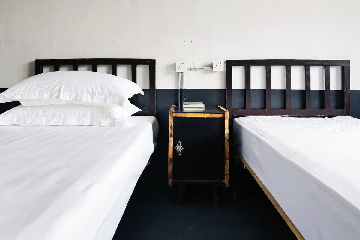 Une chambre avec deux lits simples, chacun doté de draps blancs et d’une tête de lit en bois. Une petite table de chevet en bois, sur laquelle se trouvent un livre et une lampe, est placée entre les deux lits.