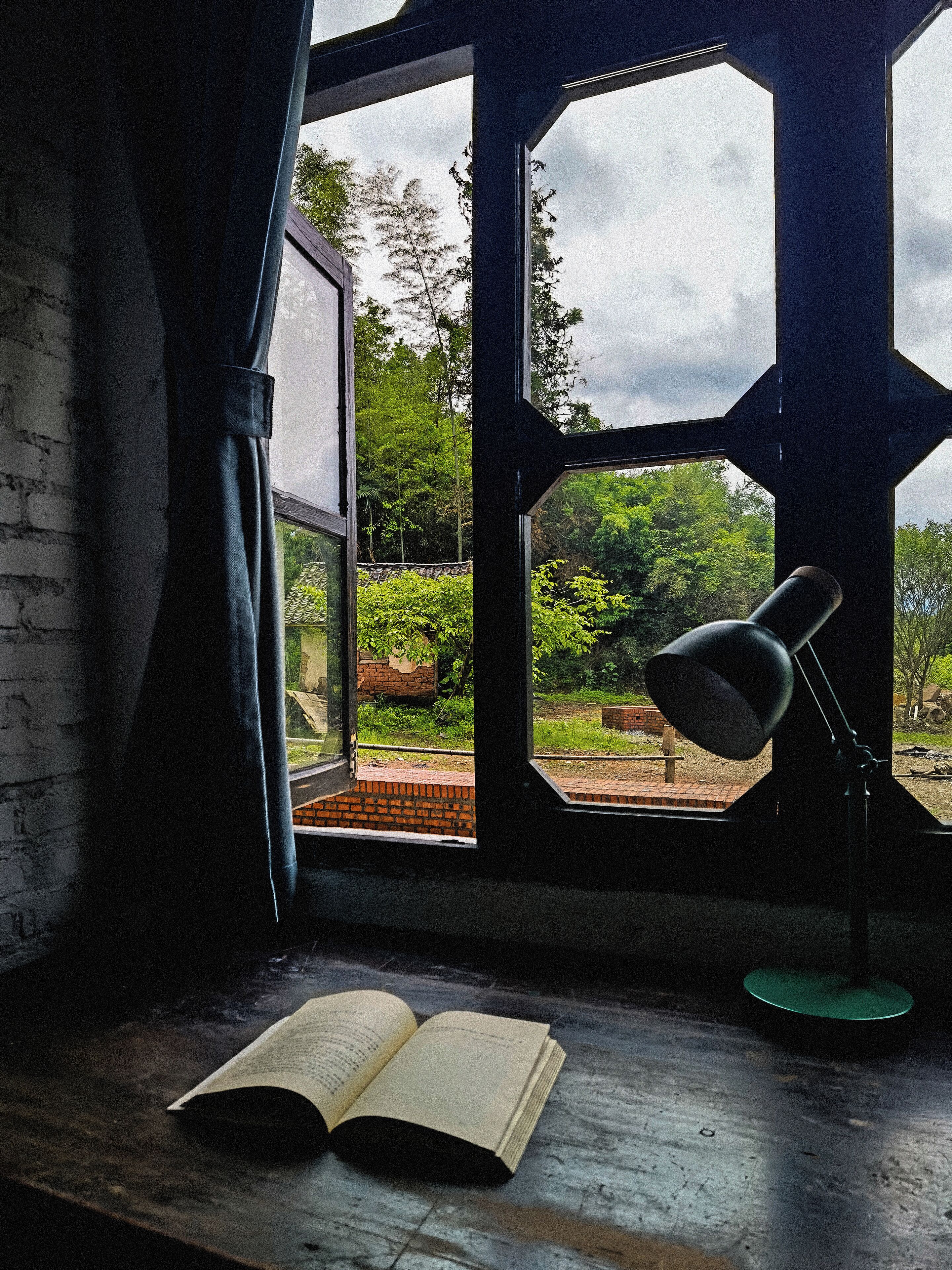 A room with a large window, a reading lamp, and an open book on a wooden table.