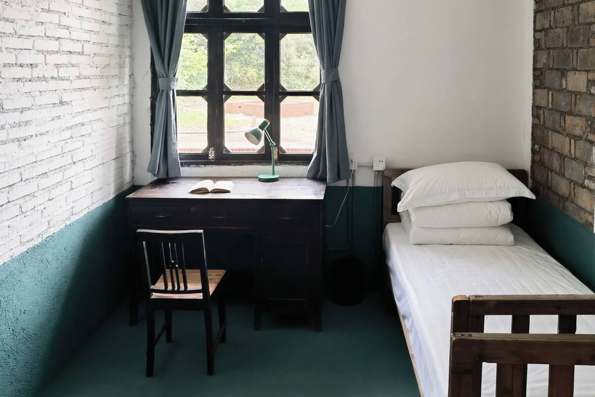 Un petit logement composé d’une seule pièce, comprenant un bureau, une chaise, un lit et une fenêtre avec des rideaux.