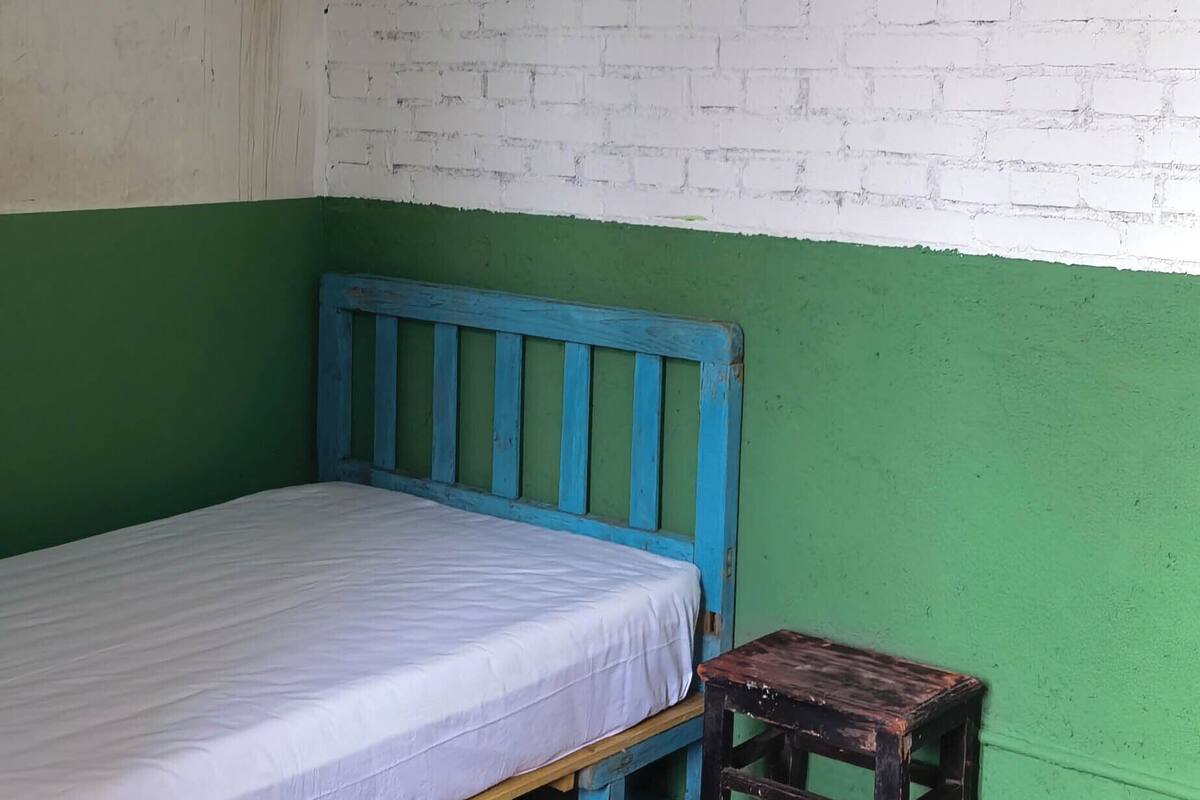 Une petite chambre avec un lit simple, une tête de lit bleue, un tabouret en bois et un mur en briques vertes et blanches.