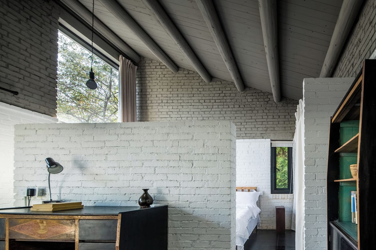 Une pièce avec un mur en briques blanches, un bureau en bois avec une lampe, un petit vase et un lit avec une literie blanche.