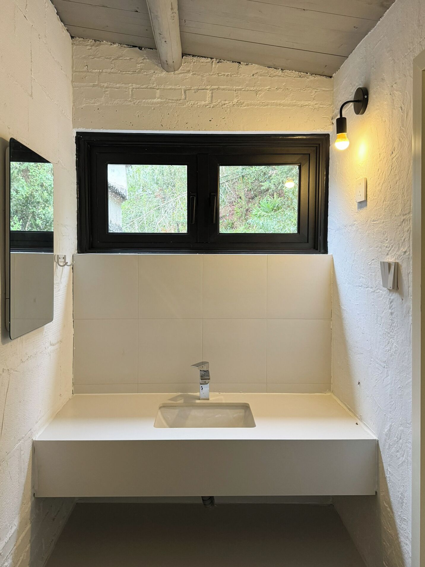 Un baño con un lavamanos blanco, una ventana sobre el espejo y una lámpara fijada a la pared.
