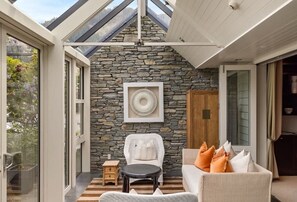 Une pièce avec un mur en pierre, une chaise blanche, un canapé avec des coussins orange, une petite table et un ventilateur.