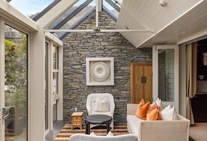 A room with a stone wall, a white chair, a sofa with orange pillows, a small table, and a fan.