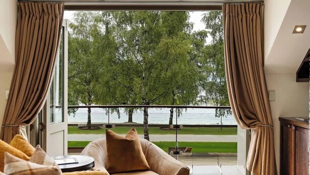 A room with a sofa, a coffee table, and a view of a lake and trees. - Creighton 1- Spacious Lakeshore Apartment (Queenstown)