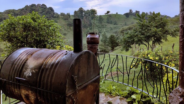 A rustic outdoor setting with a metal barrel, a chimney, and a metal fence overlooking a lush green landscape.