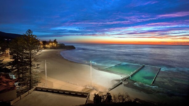 A beach with a view of the ocean and a sunset. There is a building with lights on the left side. - Austinmer On The Beach (Austinmer)