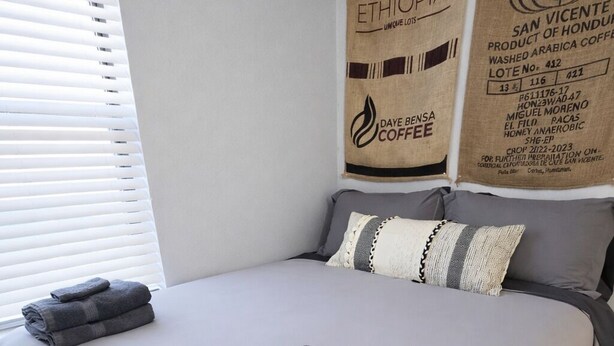 A neatly made bed with folded towels, a grey bedspread, and two pillows. There are two wall hangings with coffee-related text.