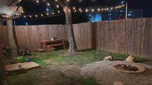A backyard with a wooden fence, a wooden table, a fire pit, and string lights.