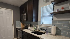 A kitchen with dark cabinets, a white dishwasher, a sink, a microwave, and a window with blinds.