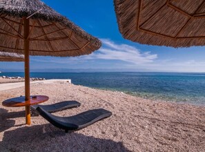 A beach with two thatched umbrellas and lounge chairs, a clear view of the sea, and a rocky shoreline. - Seaside Oasis Apartments on Island Ugljan (Preko)