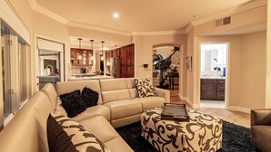 Un salon spacieux avec un canapé d’angle beige, un pouf à motifs et un tapis foncé.
