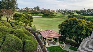 Un belvédère aux piliers de pierre et au toit en tuiles, entouré d’arbustes et d’arbres bien entretenus, offrant une vue sur un terrain de golf et, au loin, sur un quartier résidentiel.