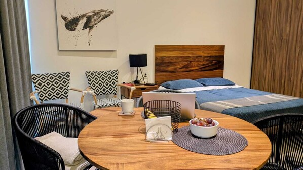 A hotel room with a wooden dining table, two black wicker chairs, a laptop, a bowl of fruit, and patterned pillows.