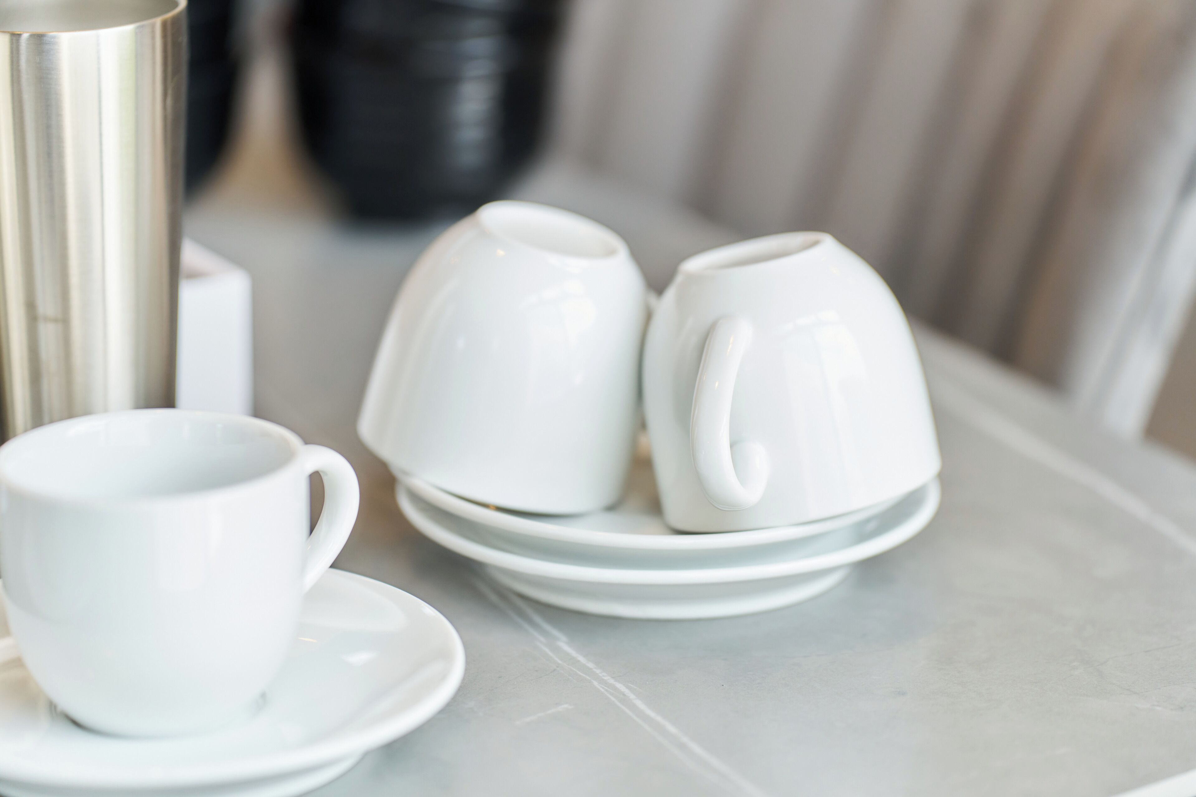 A table with white ceramic cups and saucers, a silver thermos, and a black container.