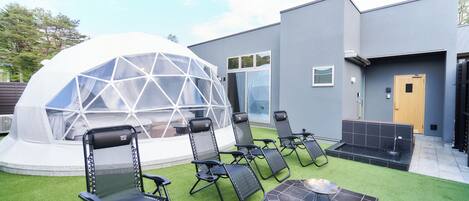 A modern outdoor seating area with lounge chairs and a hot tub, a geodesic dome structure, and a building with large windows and a door.