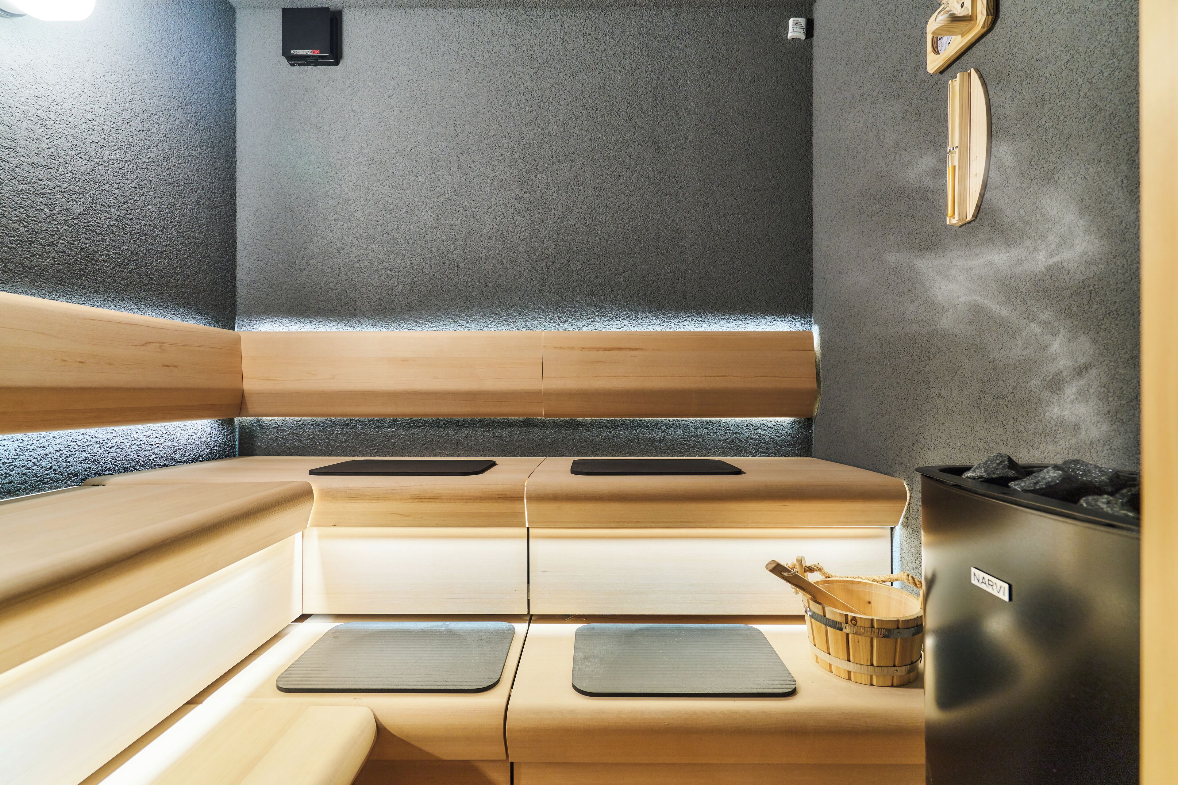 Una sala de sauna con bancos de madera, un calentador negro y un cubo de madera.