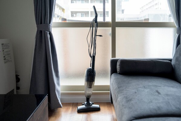 A room with a grey sofa, a standing vacuum cleaner, and a window with curtains.