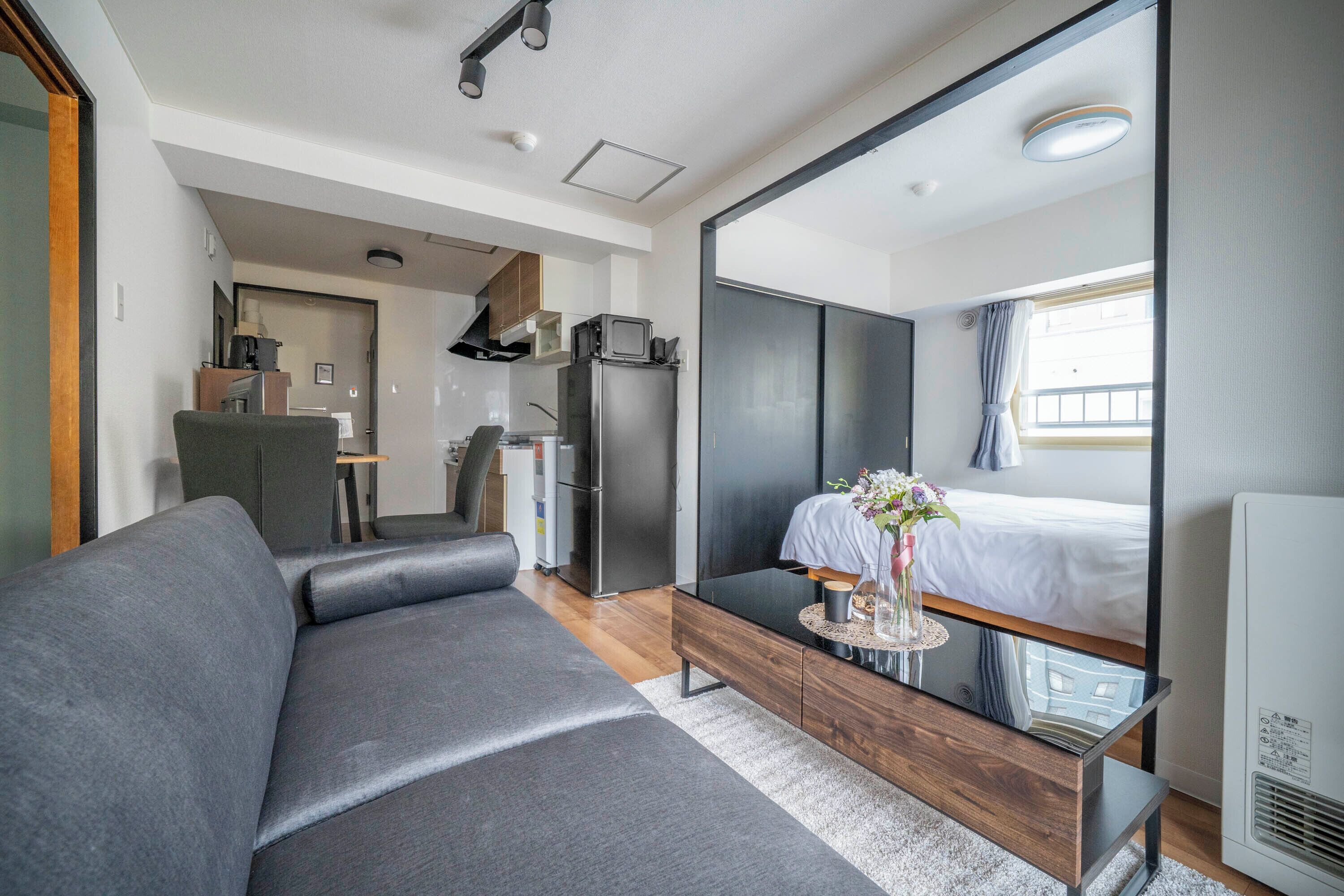 A modern hotel room with a grey sofa, a small dining table, a bed with a white duvet, and a large mirror reflecting the room.