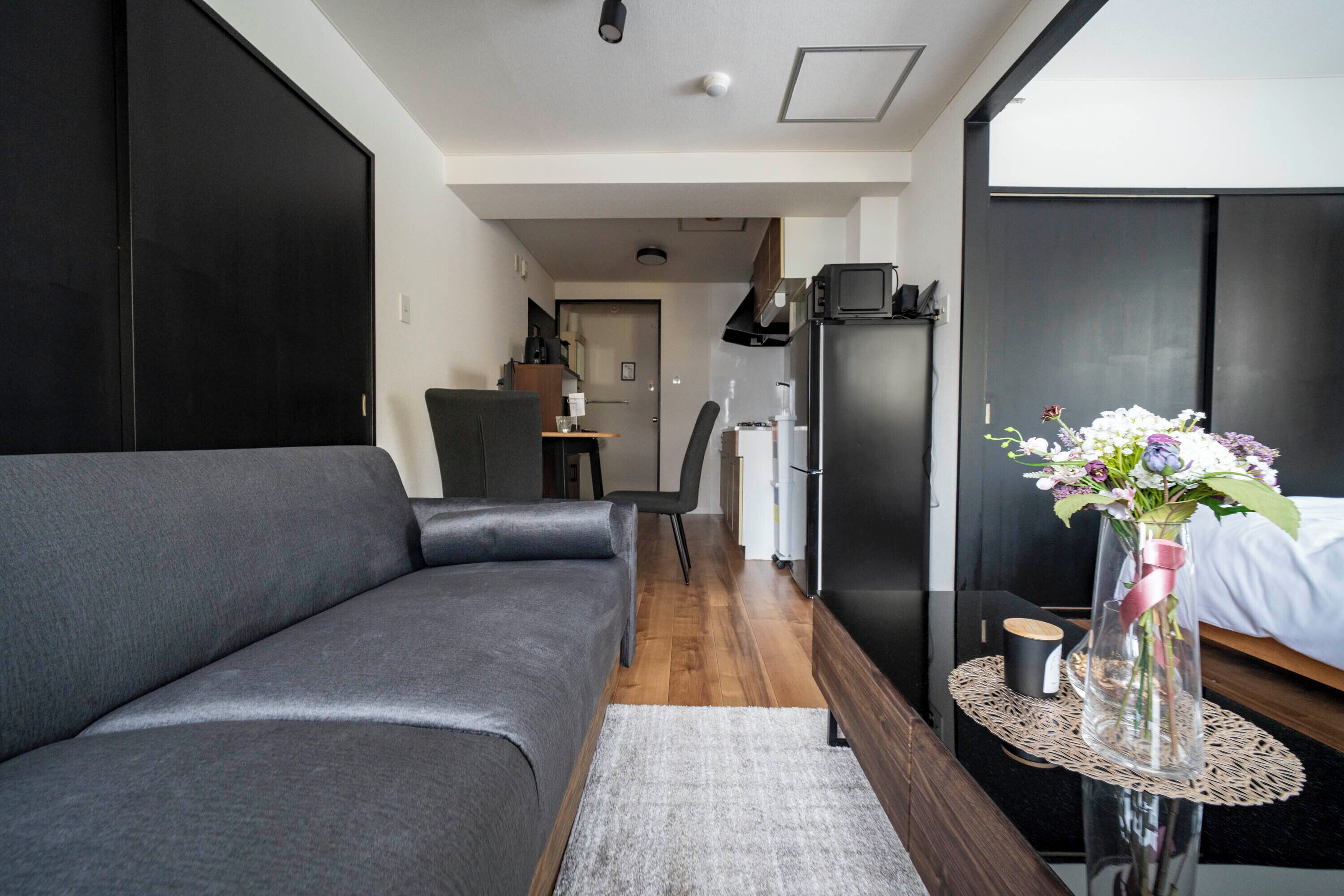 Una habitación de hotel moderna con un sofá gris, una mesita redonda con un florero y flores, una cama y una zona de kitchenette.