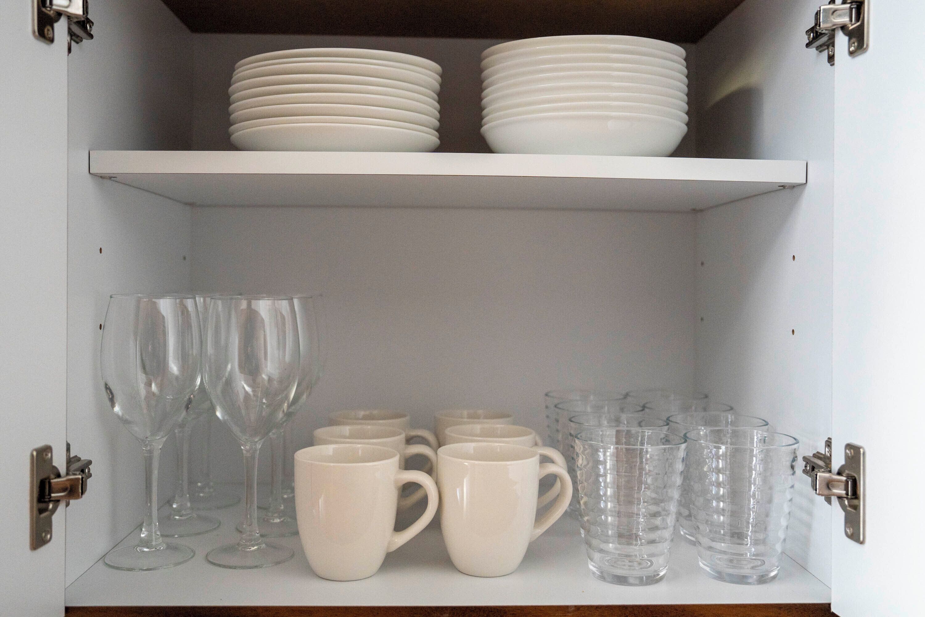 A cabinet with shelves containing white plates, clear drinking glasses, and white mugs.