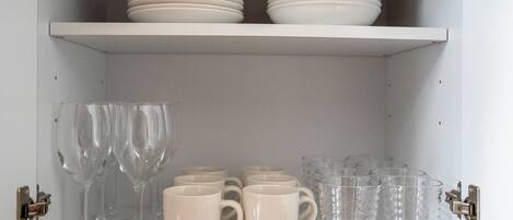 A cabinet with shelves containing white plates, clear drinking glasses, and white mugs.