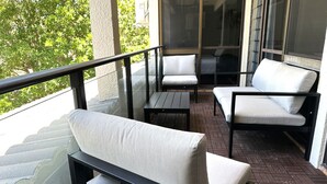 En balkong med två vita fåtöljer med mjuka kuddar och ett litet bord, ett glasräcke och en utsikt över träd och en byggnad i bakgrunden.