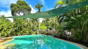 En swimmingpool med en kant som är klädd med småsten, omgiven av frodig grönska och träd.