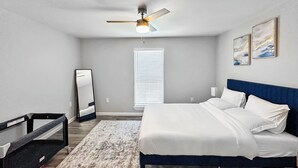 Un dormitorio con cama, ventilador de techo, espejo grande y ventana con persianas.