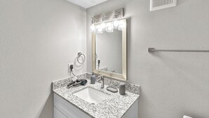 Un baño con lavamanos de encimera de granito, un espejo con luces, un toallero y un dispensador de papel higiénico.