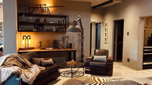 A modern living room with a sofa, a zebra-patterned ottoman, a coffee table, and a floor lamp. There is a bookshelf with decorative items and a clock. - The Citadel @ Tether&Tease, Extraordinary BDSM Location in Cape Town (Cape Town)