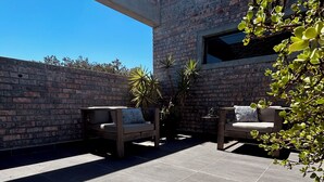 An outdoor patio with two wooden sofas, a potted plant, and a brick wall. - The Citadel @ Tether&Tease, Extraordinary BDSM Location in Cape Town (Cape Town)