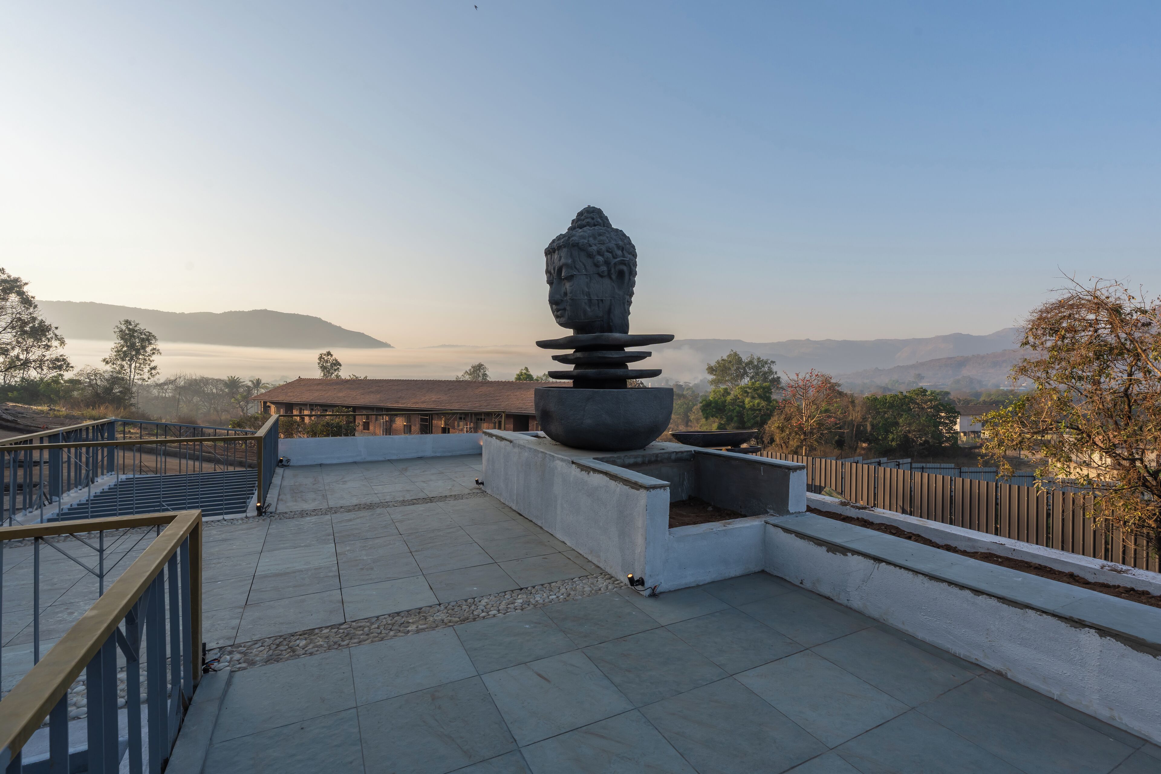 A rooftop terrace with a Buddha statue, a staircase, and a view of distant mountains and trees.